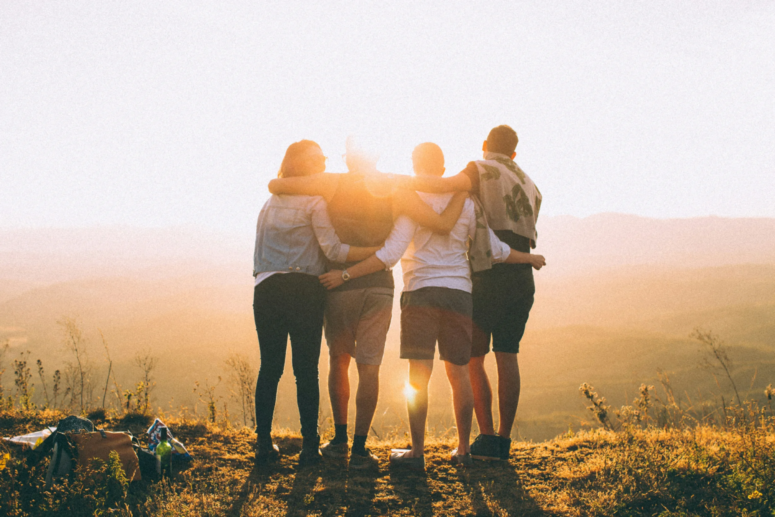 Four people standing in a field looking at the sun with their arms around each other in a team-like manner.