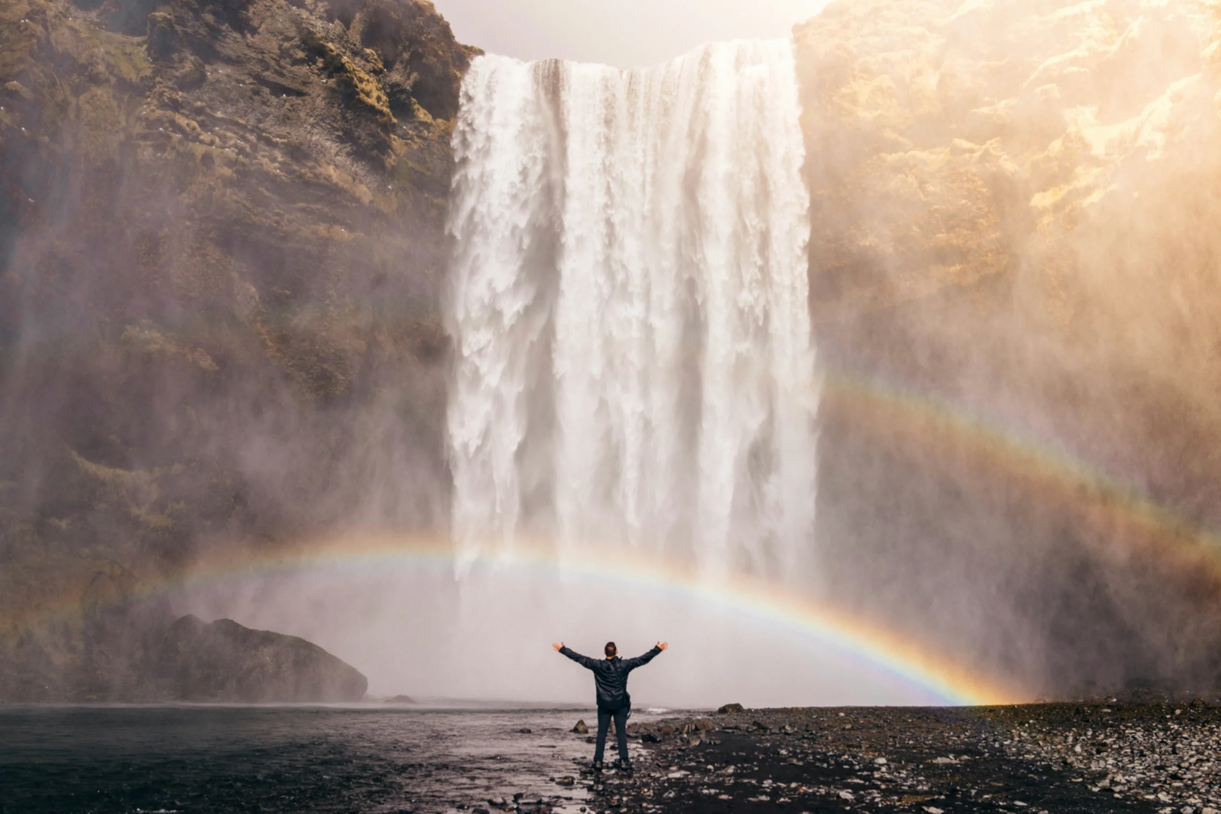 A person stands with arms outstretched in front of a large waterfall, framed by two rainbows in a misty landscape.