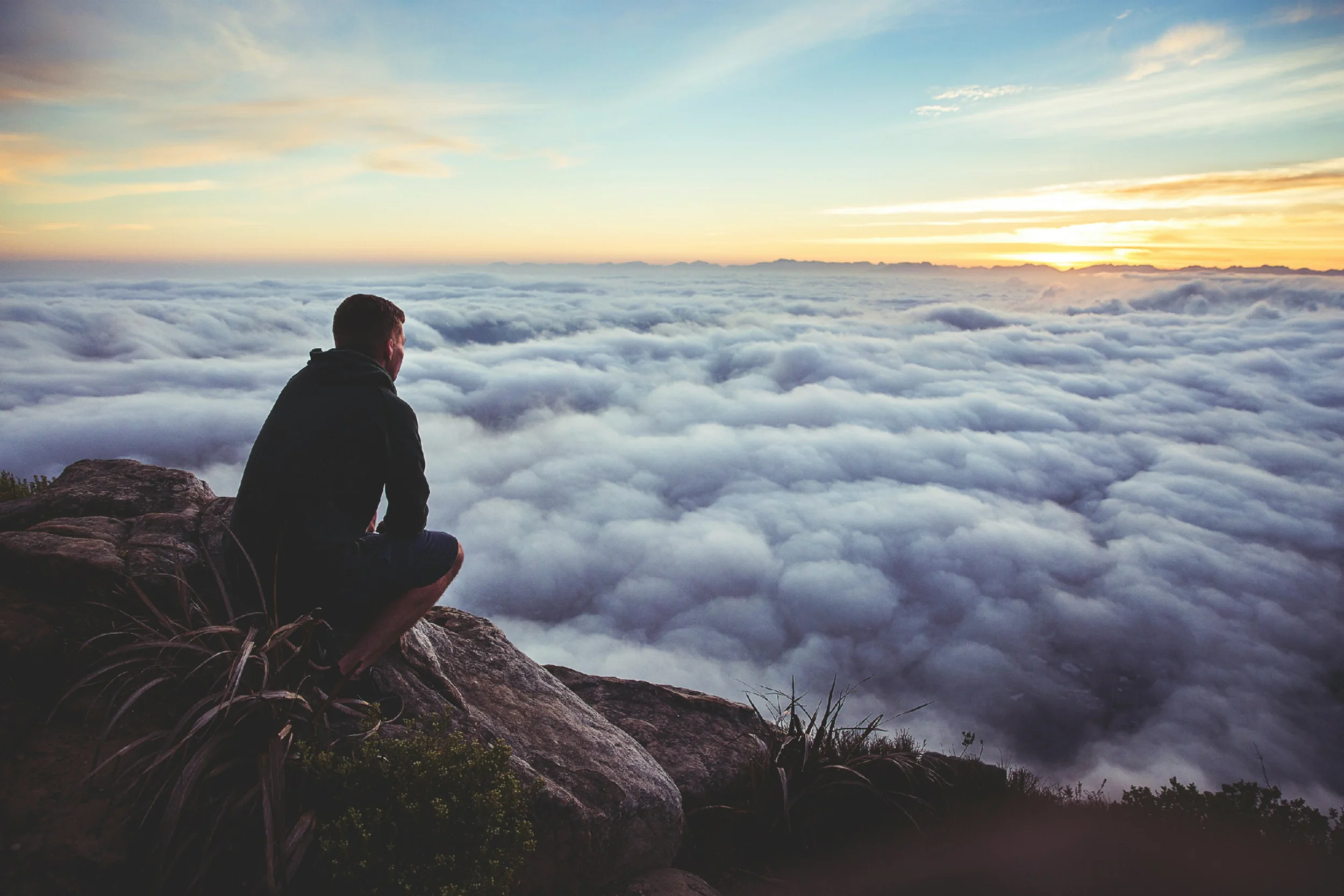 Man sits on a rock looking over a sea of clouds stretching out below him.