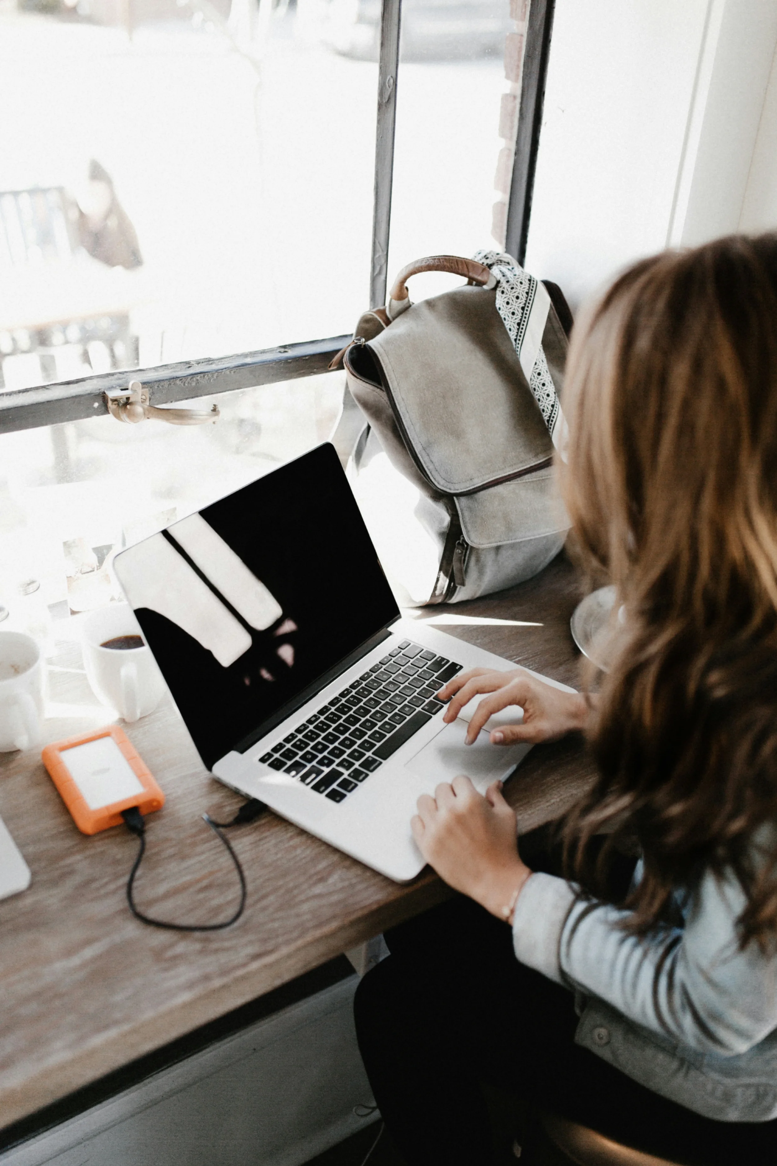 Woman working on a laptop sitting inside at a wooden bench next to a window.