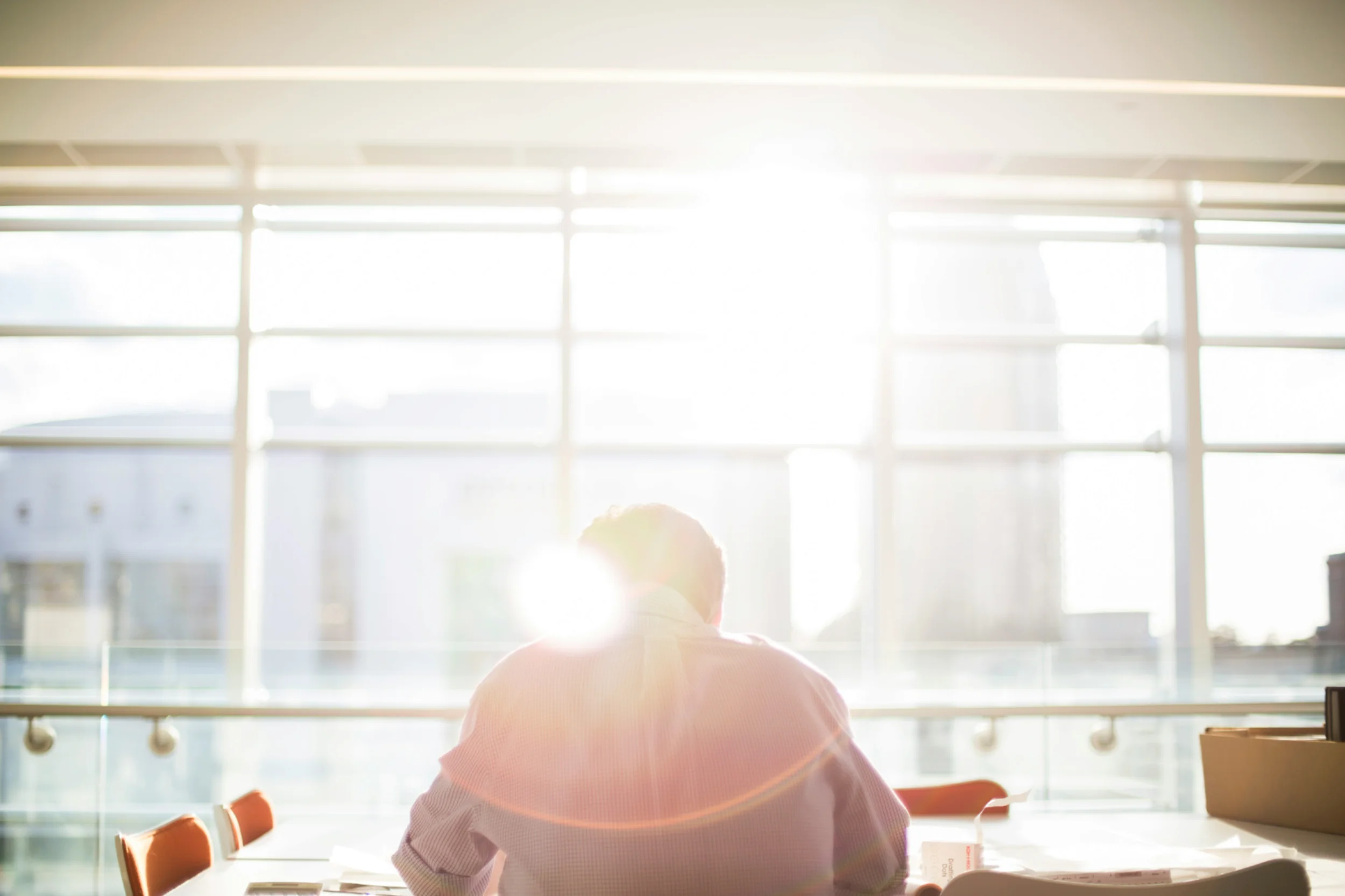 Man in a shirt sitting alone at an office meeting table facing a window through which the sun is shining.