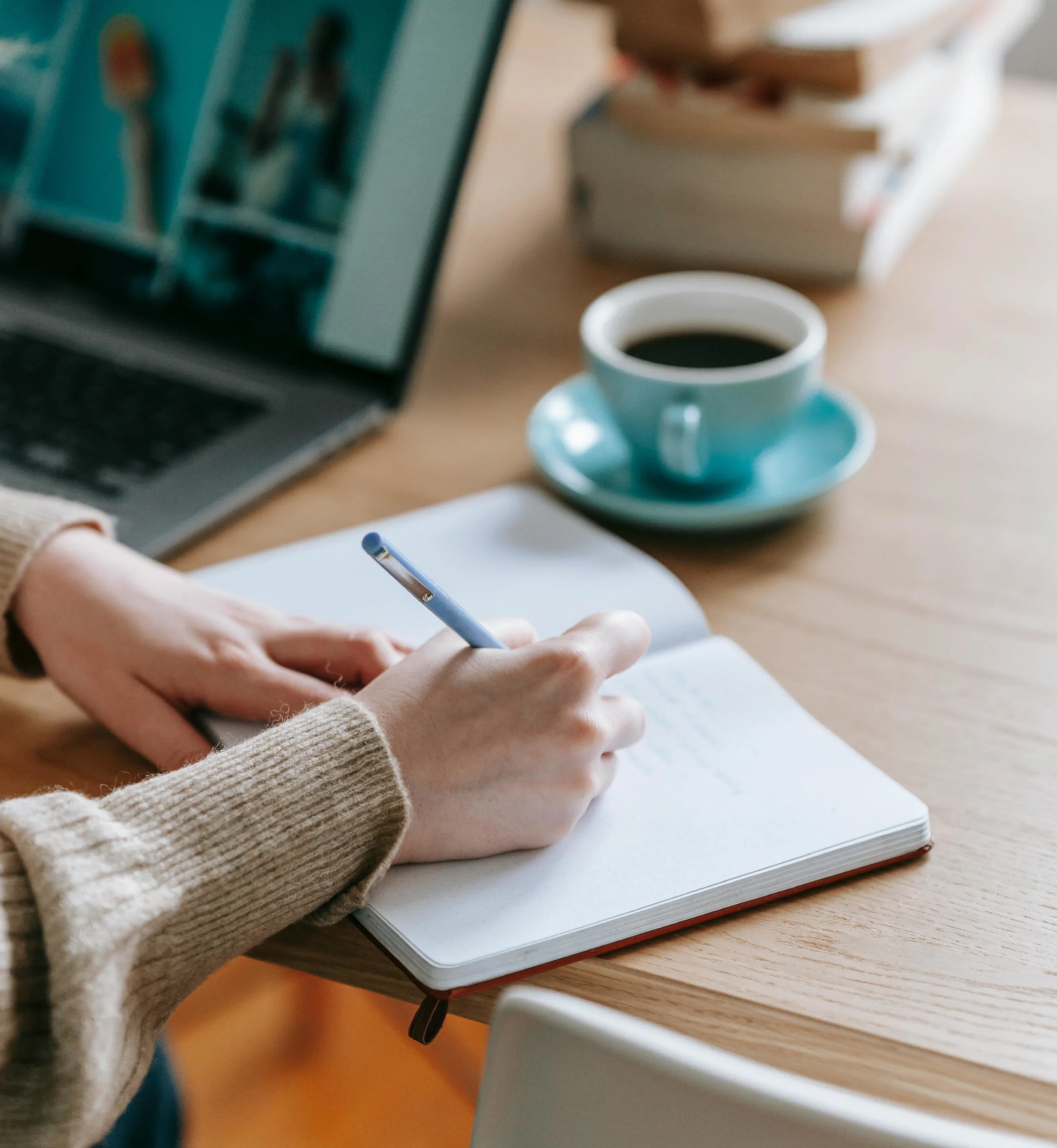 Woman's hand writing on a notepad next to a laptop and cup of coffee