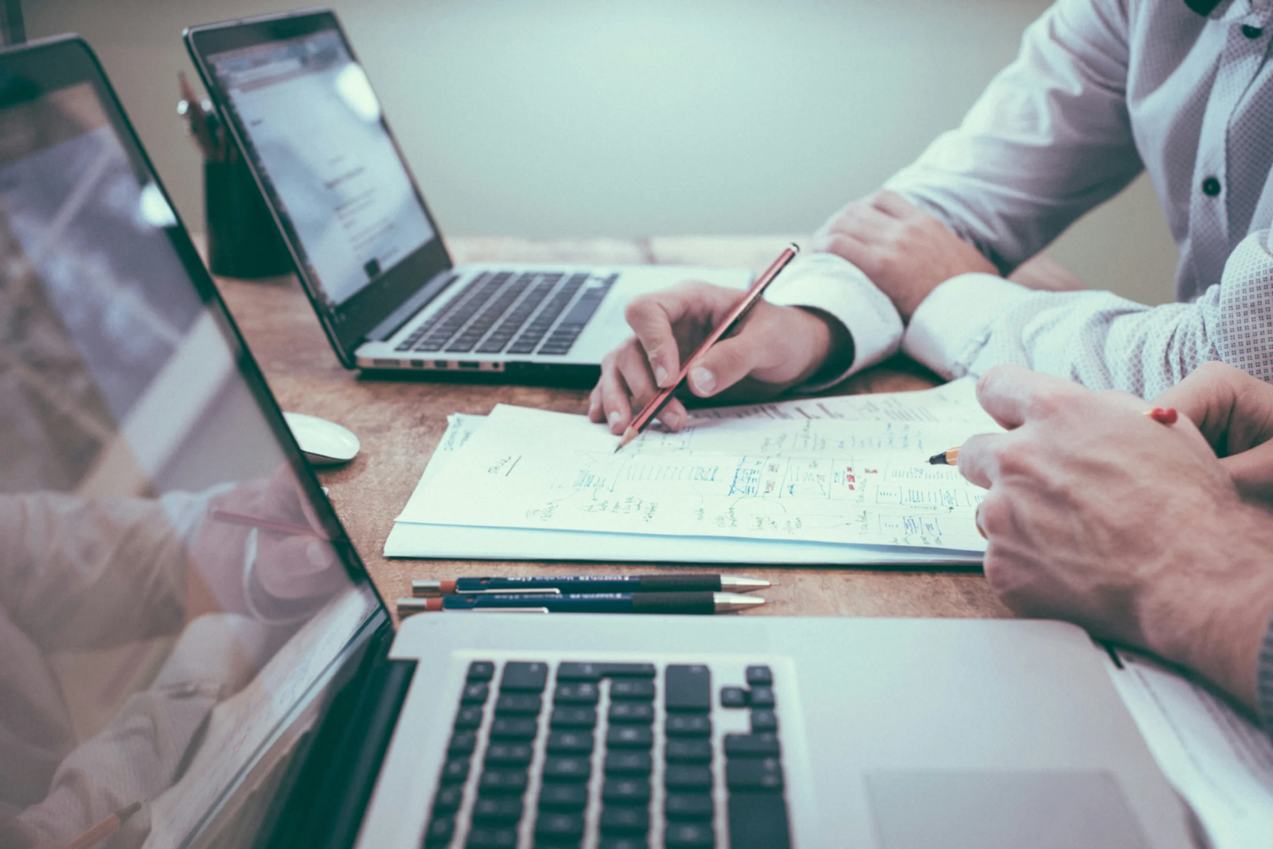Man's hand holding a pencil over a notepad sitting between two open laptops.